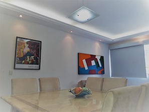 Sala de conferência com mesa de vidro, cadeiras bege, duas pinturas abstratas, iluminação embutida no teto e janela com persianas.