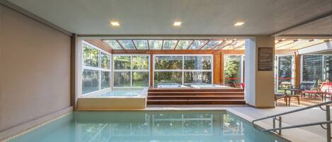 An indoor swimming pool with clear glass walls, steps leading to a seating area, and a view of greenery outside.