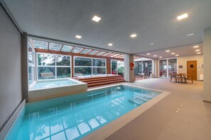 An indoor swimming pool with clear glass walls, a tiled floor, and a seating area with steps leading to the pool. - Hotel Laghetto Stilo Borges (Gramado)