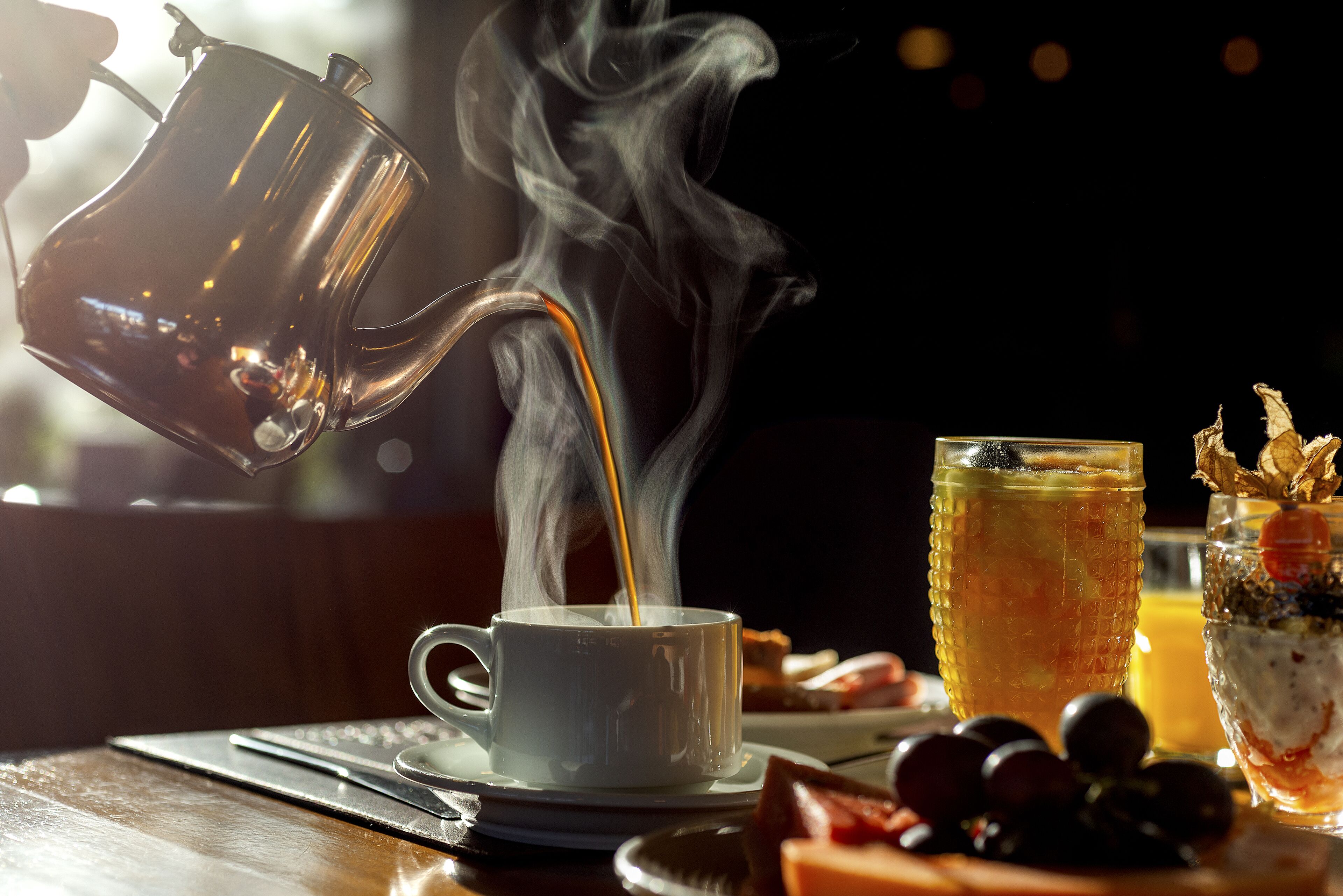 Una cafetera vertiendo en una taza, un vaso de té helado y un plato con fruta sobre una mesa de madera.
