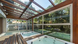 Area benessere con due vasche idromassaggio sotto una copertura in vetro, circondata da un solarium in legno e vista su un giardino.