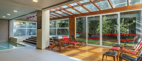 A modern indoor pool area with a glass ceiling, wooden flooring, and multiple lounge chairs.