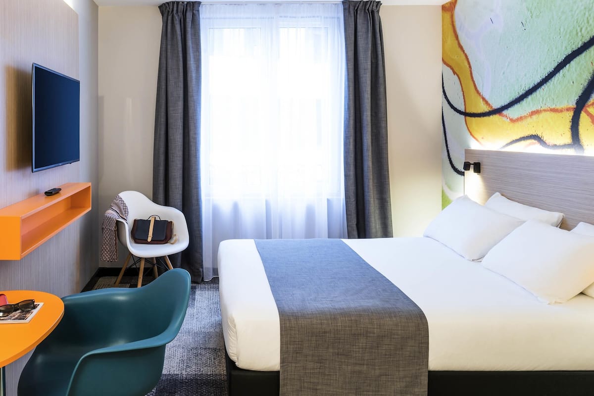 Habitación de hotel con una cama, un televisor montado en la pared, un escritorio con una silla y un cuadro grande en la pared.
