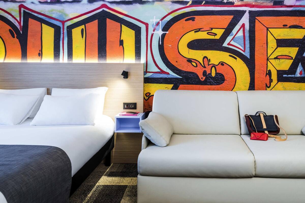 Una habitación de hotel con una cama, un sofá, una mesita de noche y una pared con grafitis de colores.