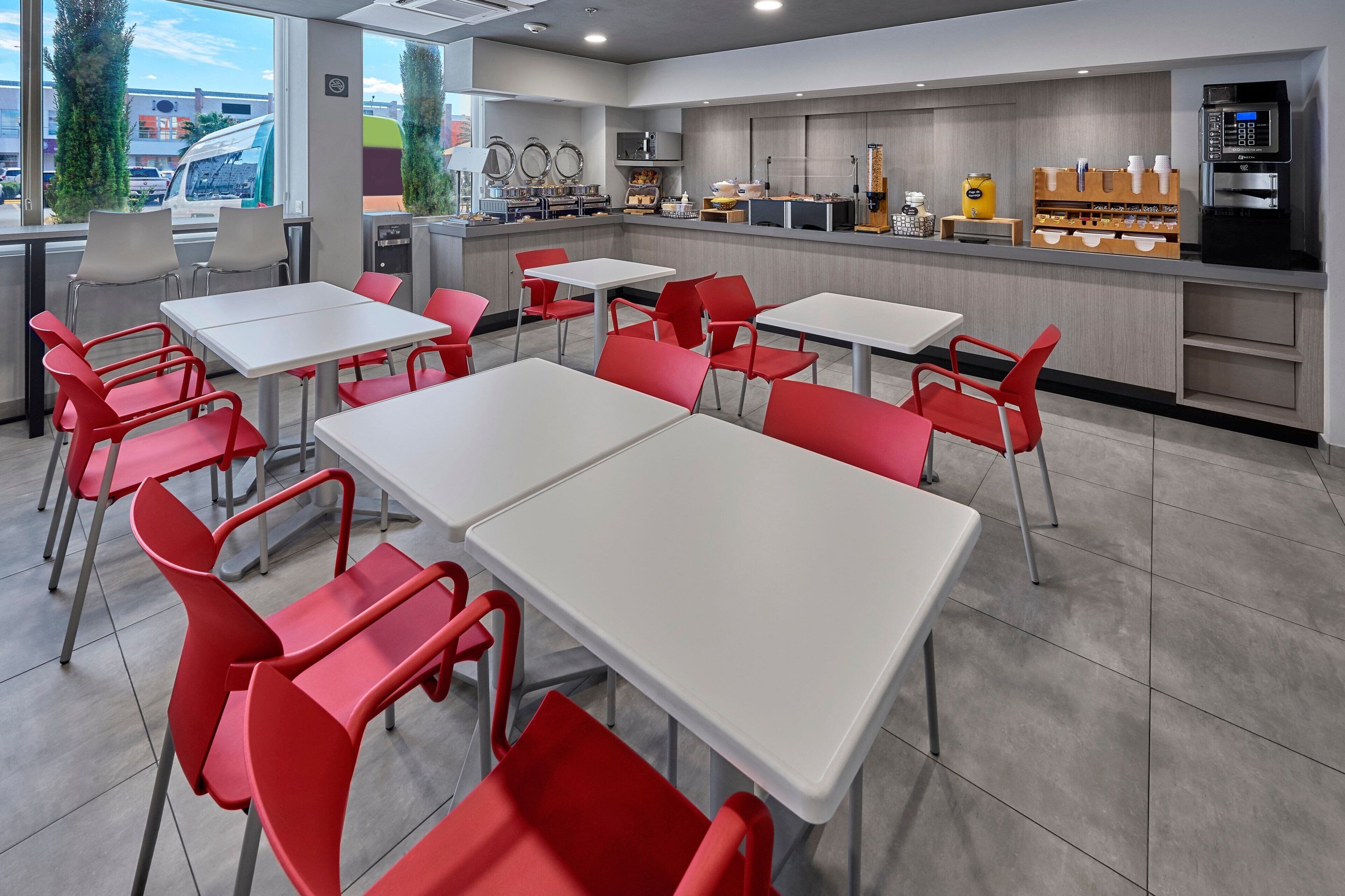 Ein modernes Restaurant mit weißen Tischen und roten Stühlen, einem Küchenbereich und einer Kaffeemaschine.