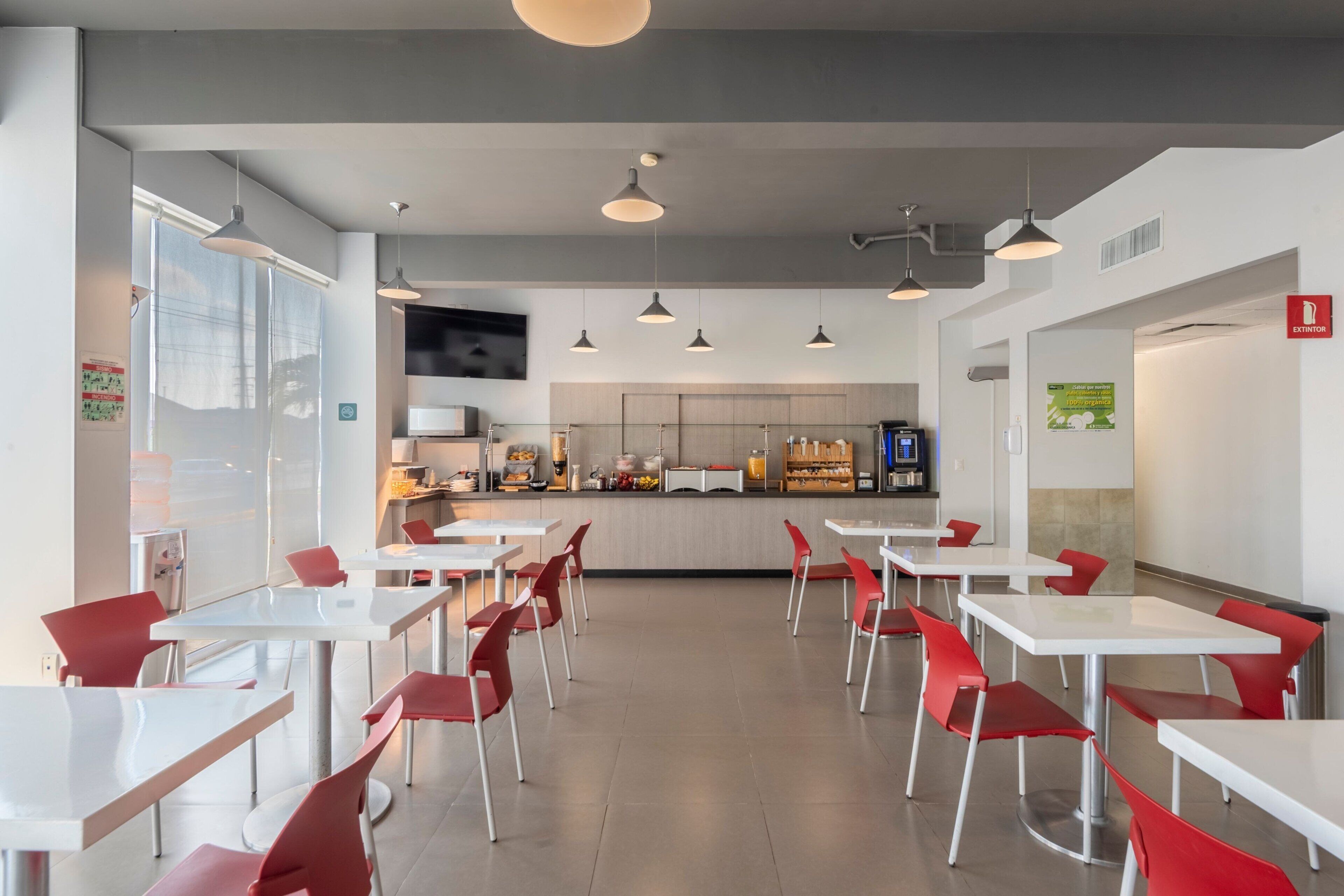 Un comedor moderno con mesas blancas y sillas rojas, una cocina al fondo e iluminación colgante.