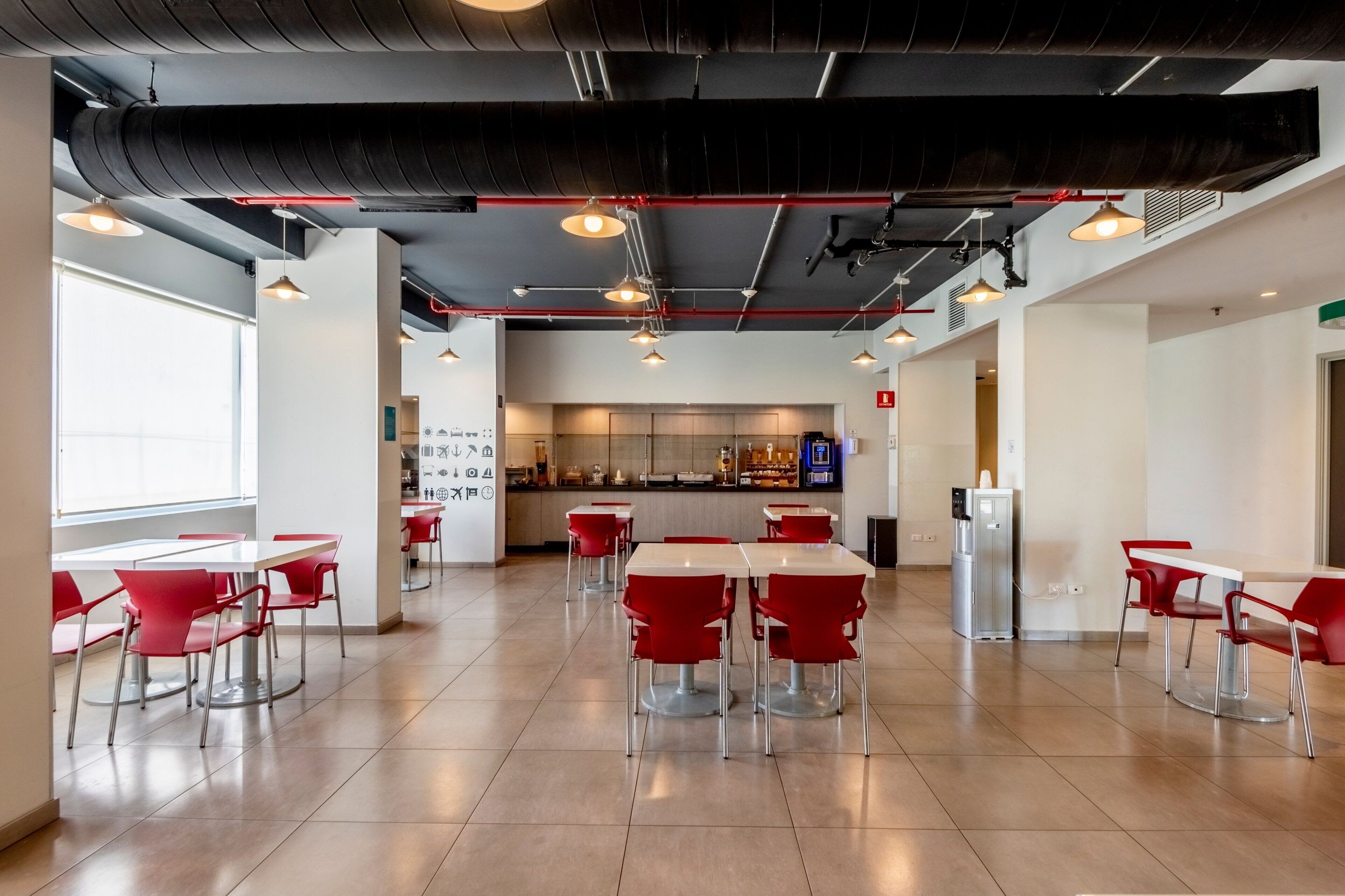 A modern cafeteria with red chairs and white tables, featuring a central counter with food display and a refrigerator.