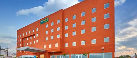 A modern hotel with a red facade, multiple floors, and a parking lot with a van in front.