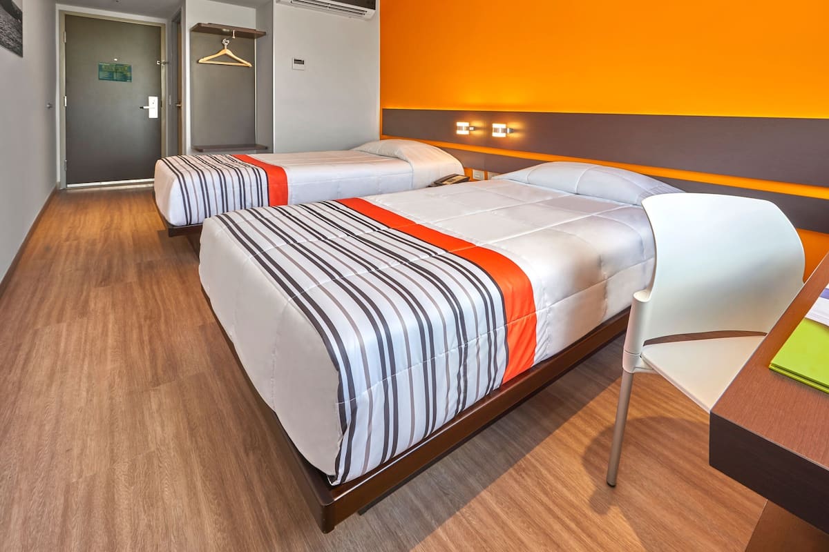 A hotel room with two beds, a wooden floor, a desk with a chair, and a wall-mounted light fixture.