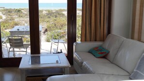A room with a white sofa, a small table, and a view of the beach and ocean. - Escape Beach House (Bergrivier)