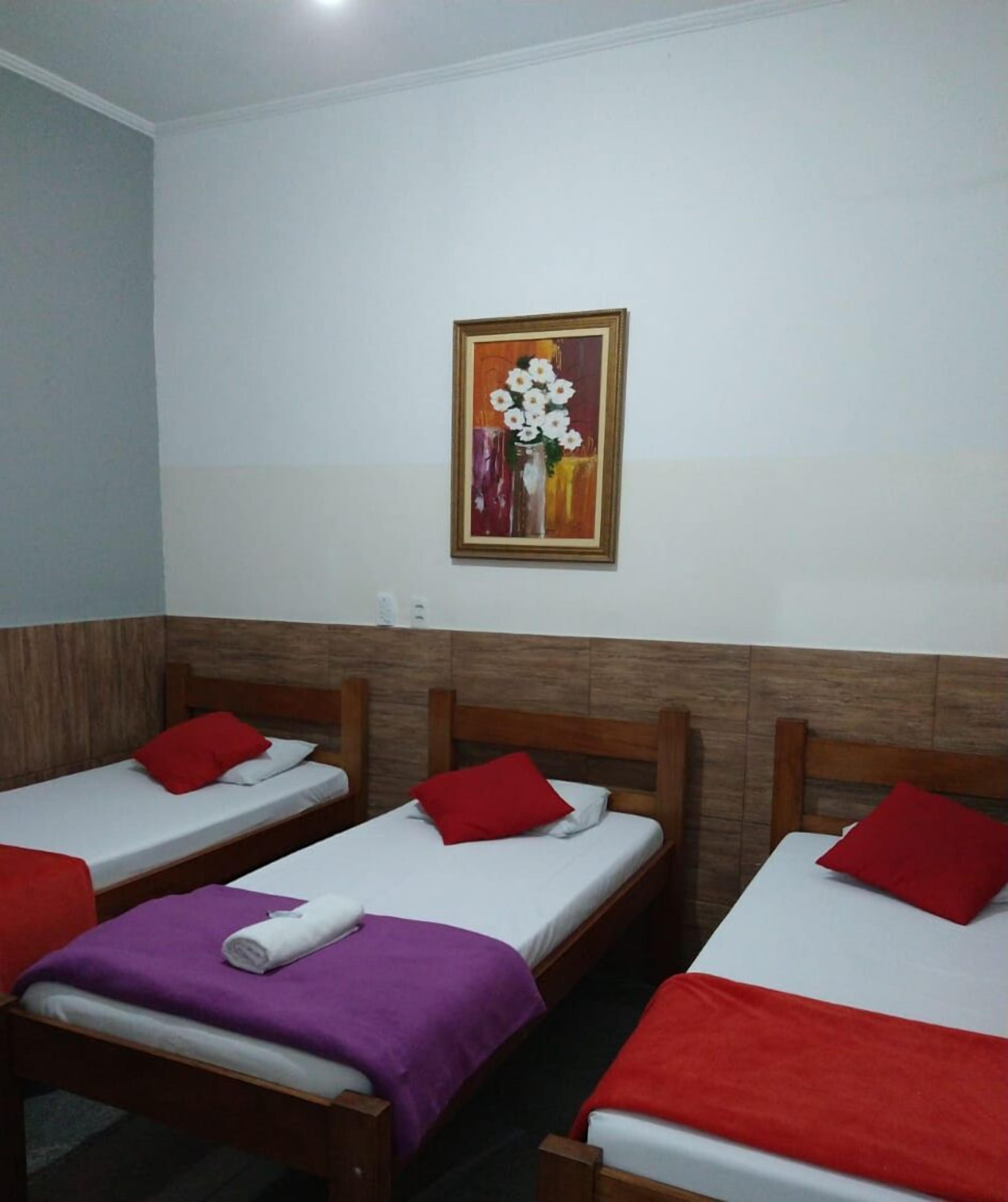 A hotel room with three single beds, each with a red pillow and a purple blanket. There is a painting of flowers on the wall above the beds.