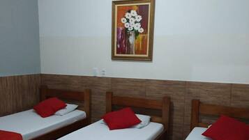 A hotel room with three single beds, each with a red pillow and a purple blanket. There is a painting of flowers on the wall above the beds.