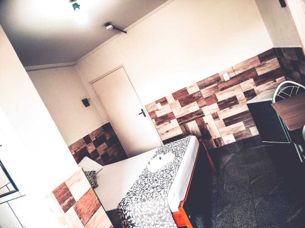 Een kleine, compacte kamer met een bed, een bureau en een stoel. Er is een deur en een muur met een visgraatmotief.