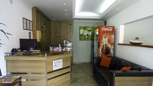 Une salle d’attente avec un bureau en bois, un ordinateur et un panneau indiquant que le port du masque est obligatoire. On y trouve un canapé avec un coussin orange et une affiche publicitaire de Coca-Cola au mur.