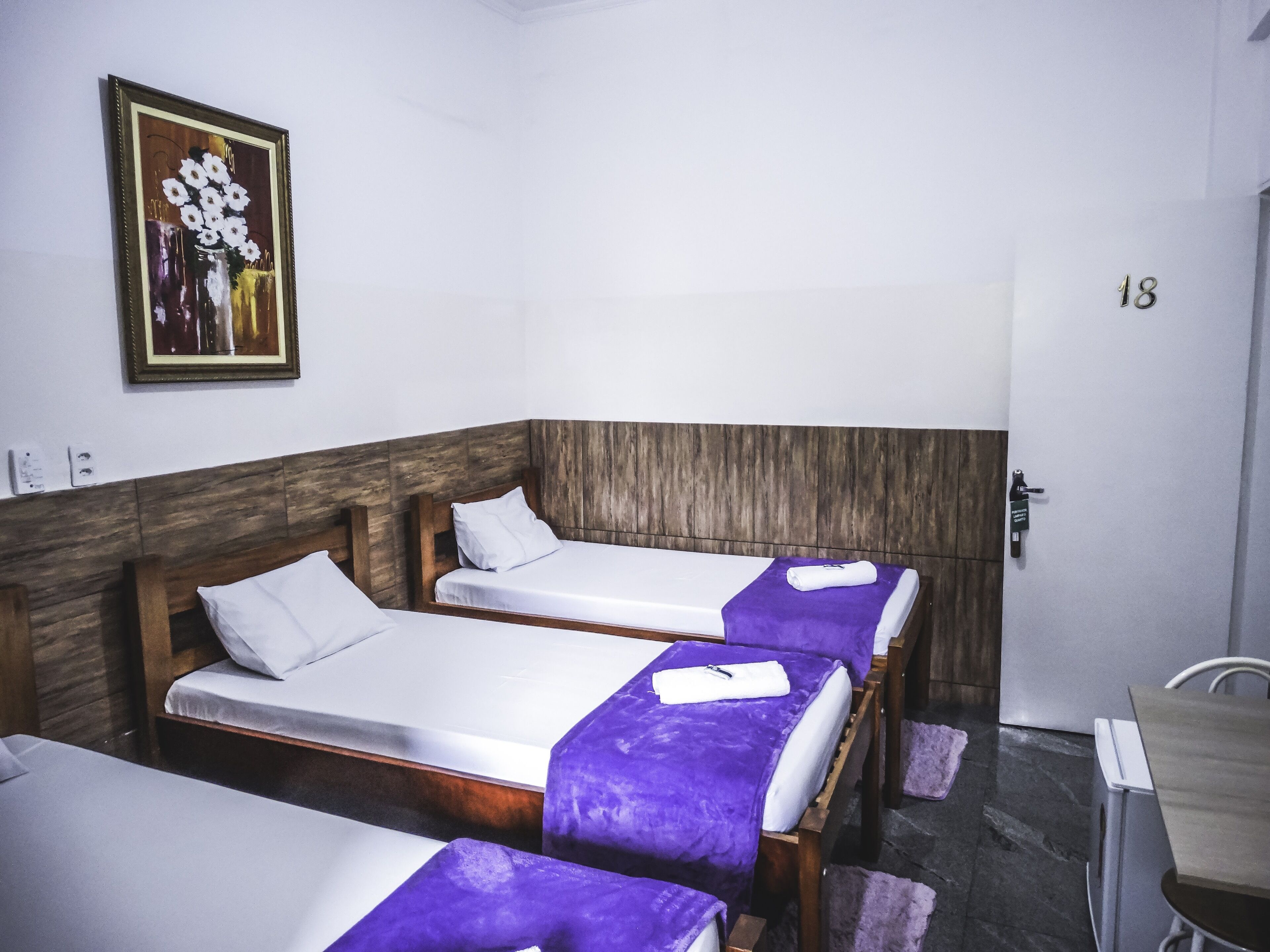A hotel room with three single beds, a wooden headboard, a door with the number 18, and a painting on the wall.