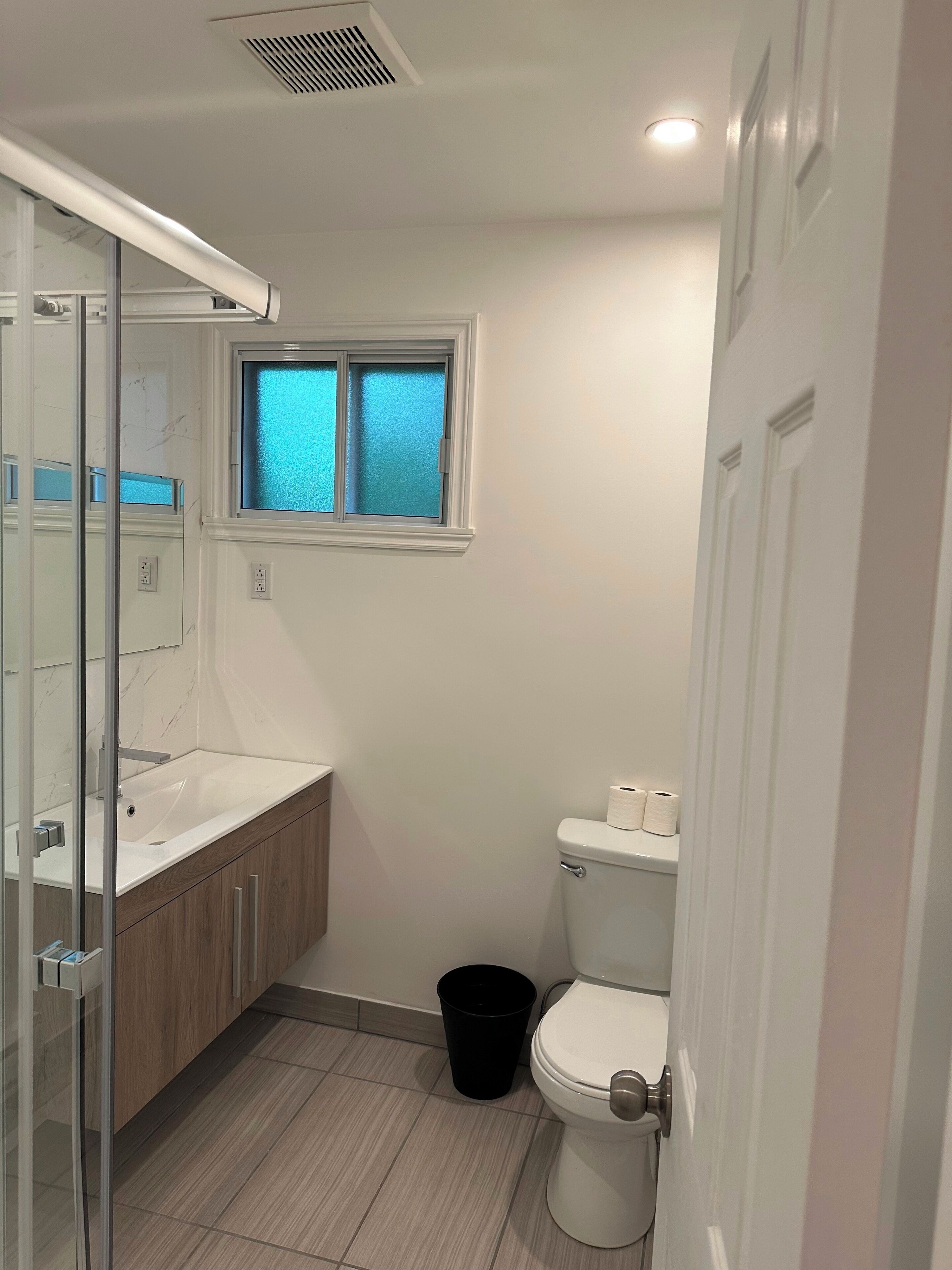 A bathroom with a glass shower enclosure, a white sink, a toilet, and a window with frosted glass.