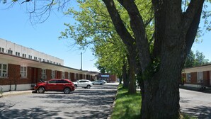 A parking lot with a red car and a white car parked in front of a building with a brick facade. - Motel Chez Nous (Laval)