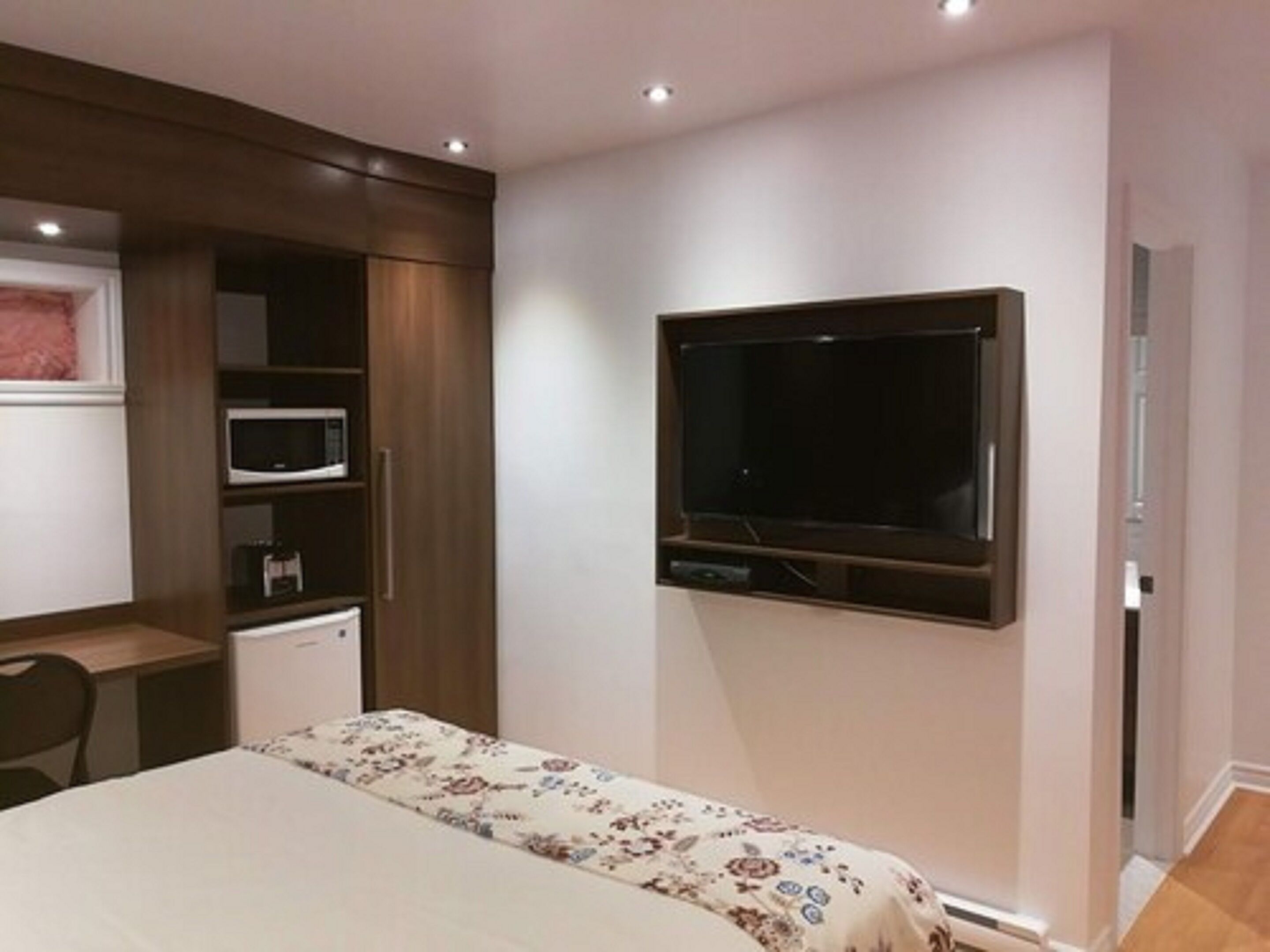 A bedroom with a bed, a TV mounted on the wall, a microwave, and a small desk with a chair.