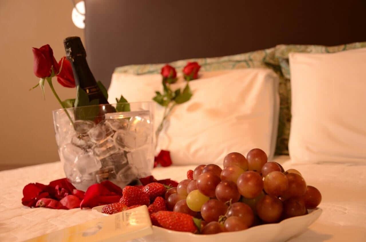 Uma cama com uma garrafa de champanhe, gelo, morangos e uvas sobre um lençol branco.