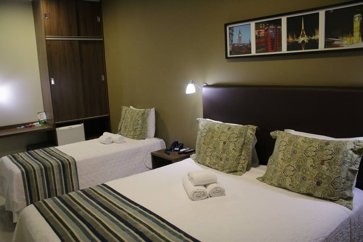 A hotel room with two beds, each with a pillow and a rolled towel. There is a nightstand with a lamp and a phone.