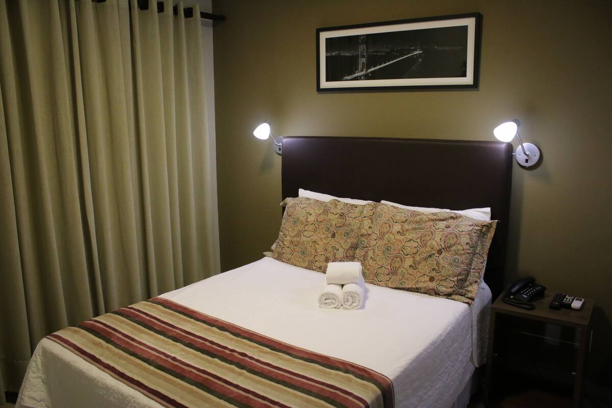 A hotel room with a bed, a nightstand with a phone, and a framed picture on the wall.