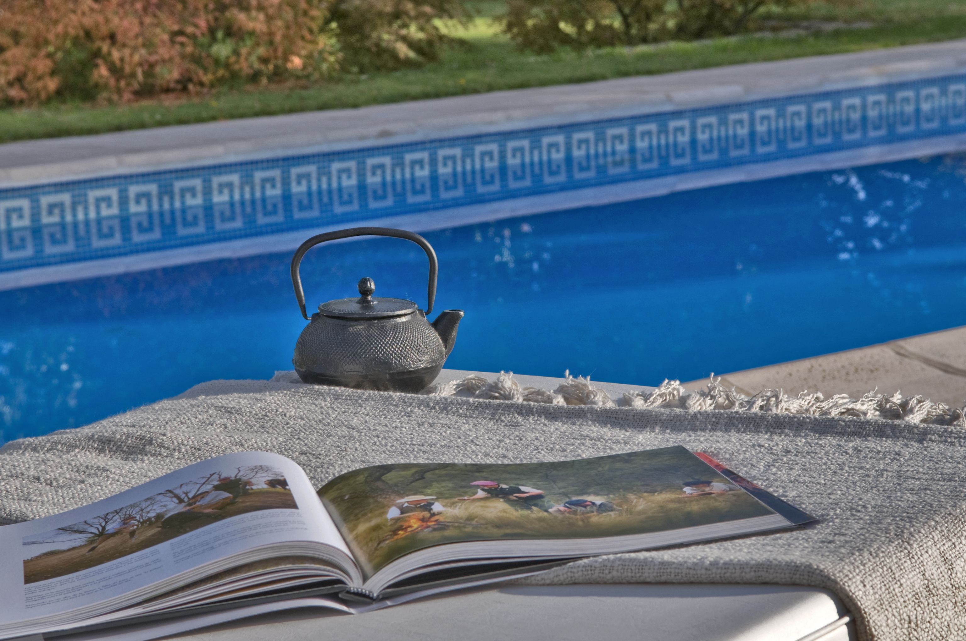 Un libro y una tetera sobre una superficie texturizada junto a una piscina con borde decorativo.