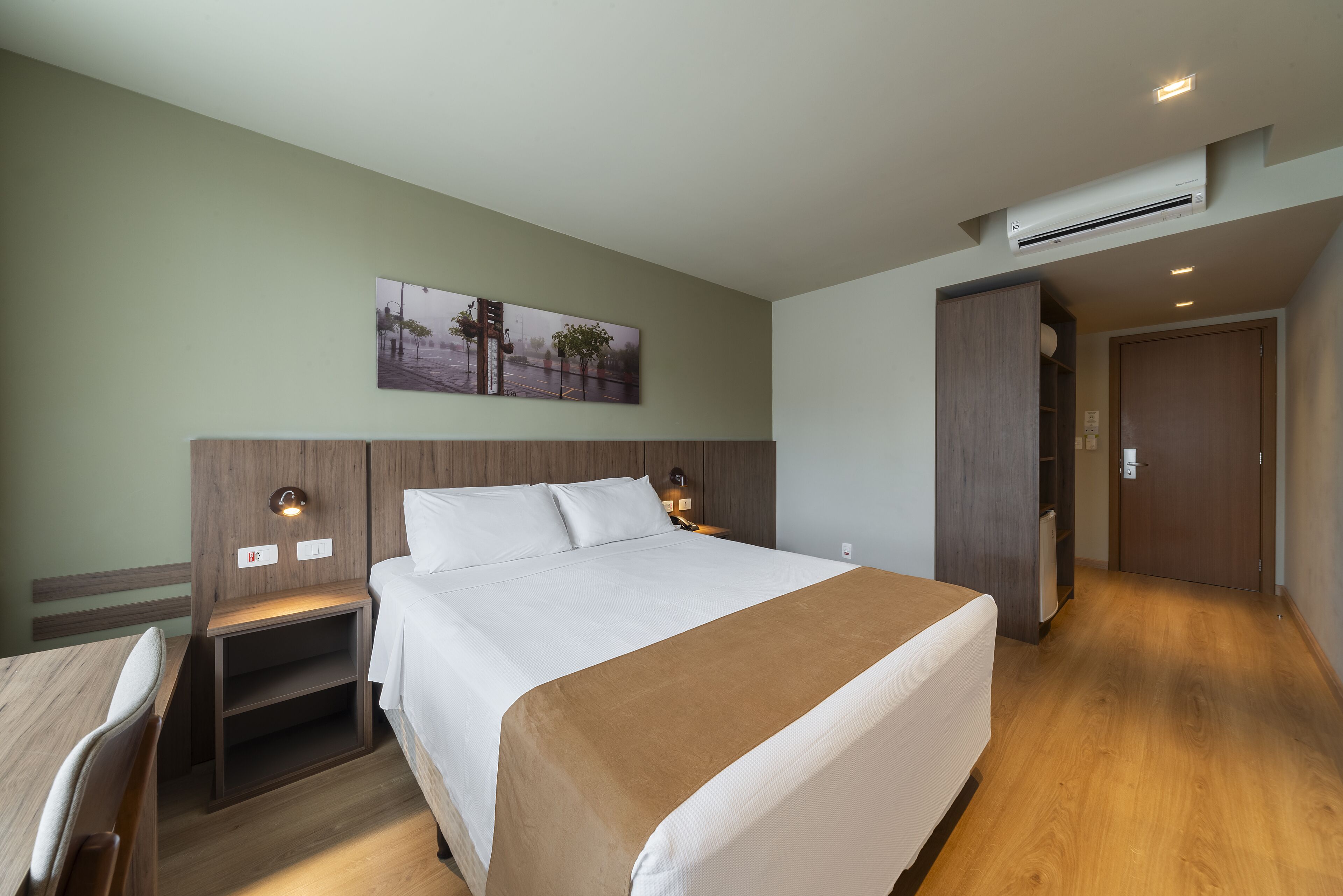 A hotel room with a large bed, a wooden headboard, a nightstand, a desk, and a wall-mounted air conditioner.