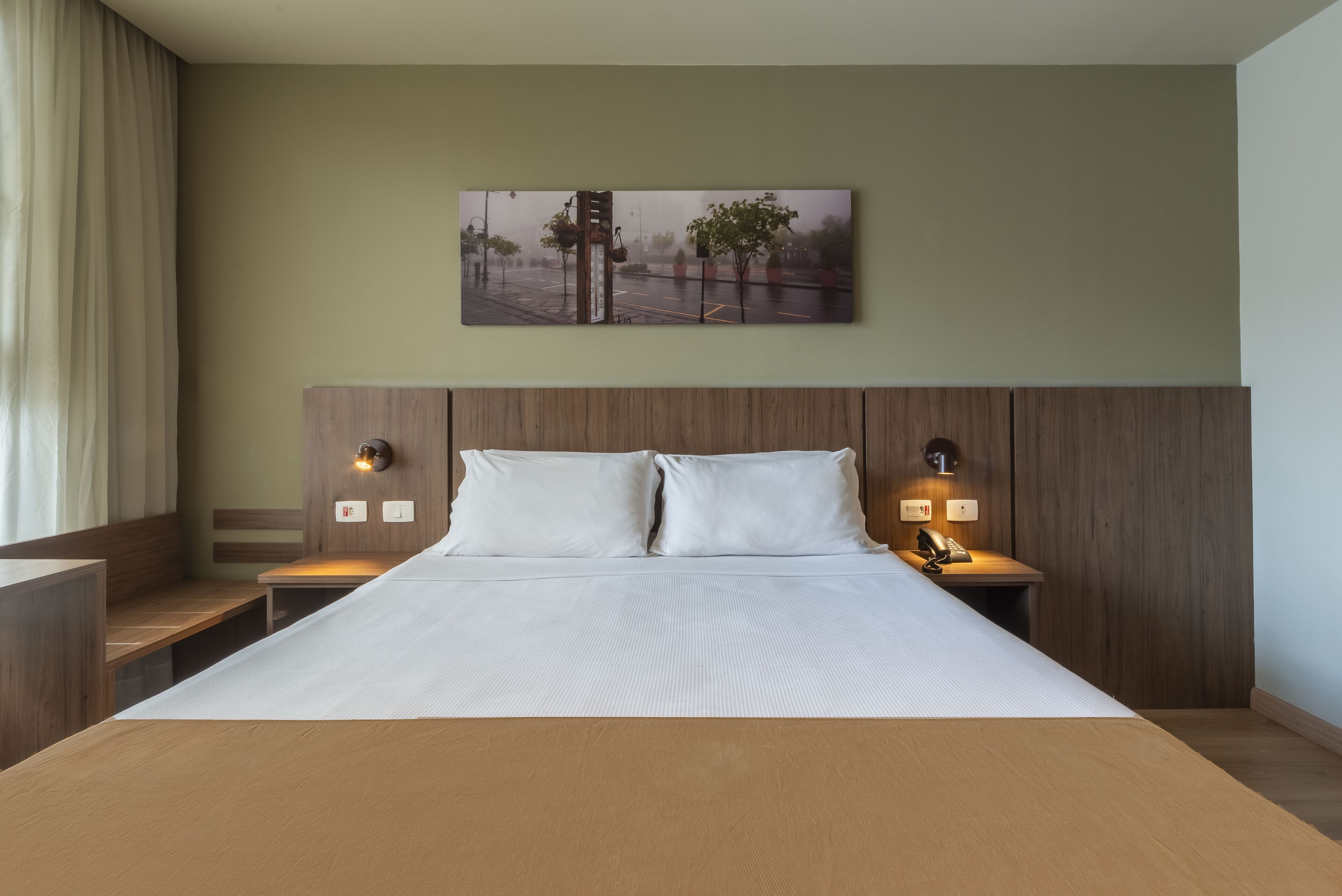 A hotel room with a large bed, wooden headboard, two bedside tables with lamps, and a wall-mounted painting.