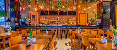 Un restaurante con mesas y sillas de madera, una zona de bar con estantes llenos de botellas y banderas de Brasil colgadas.