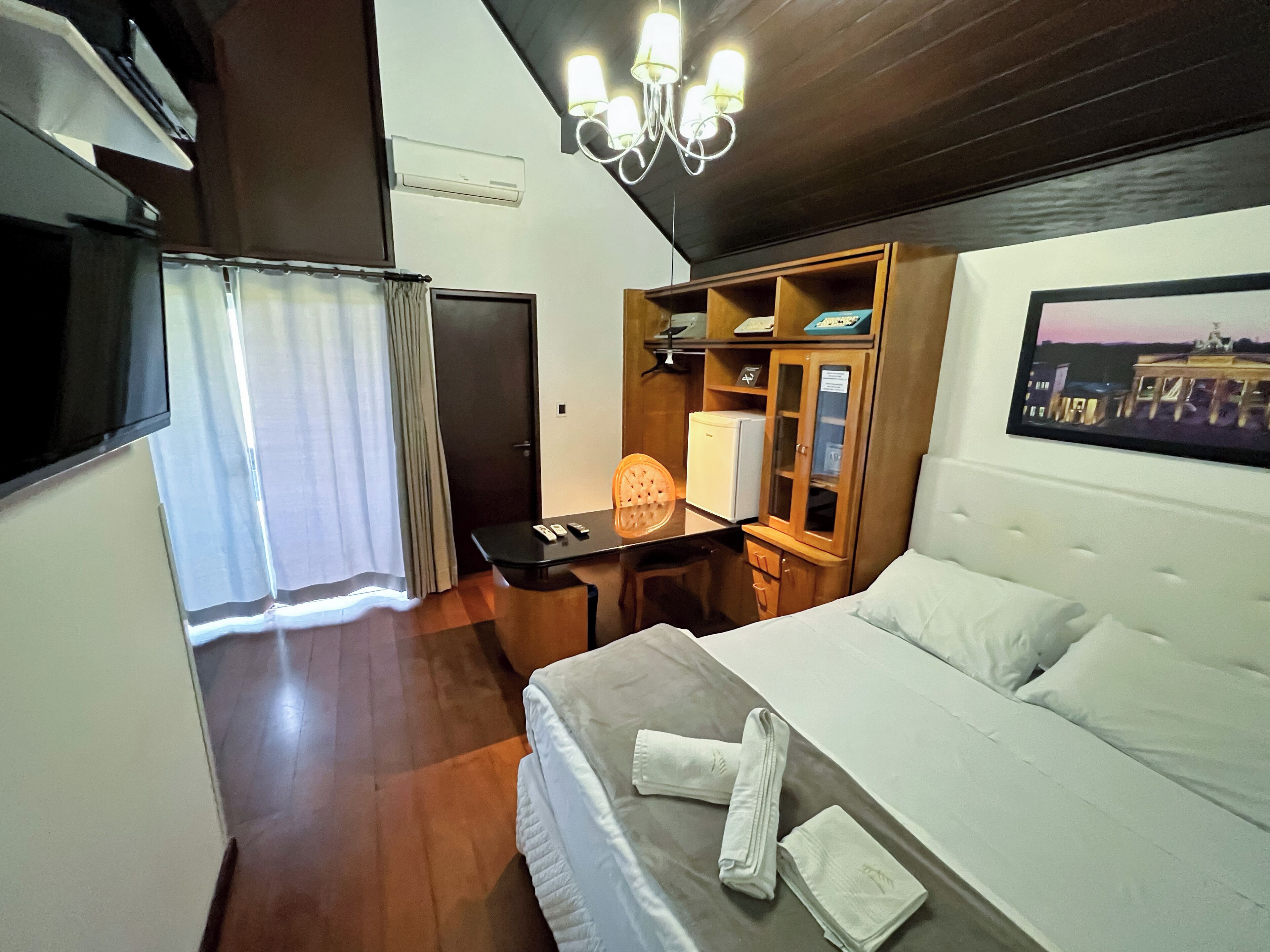 Habitación de hotel con cama, escritorio con silla, armario, televisor y candelabro.