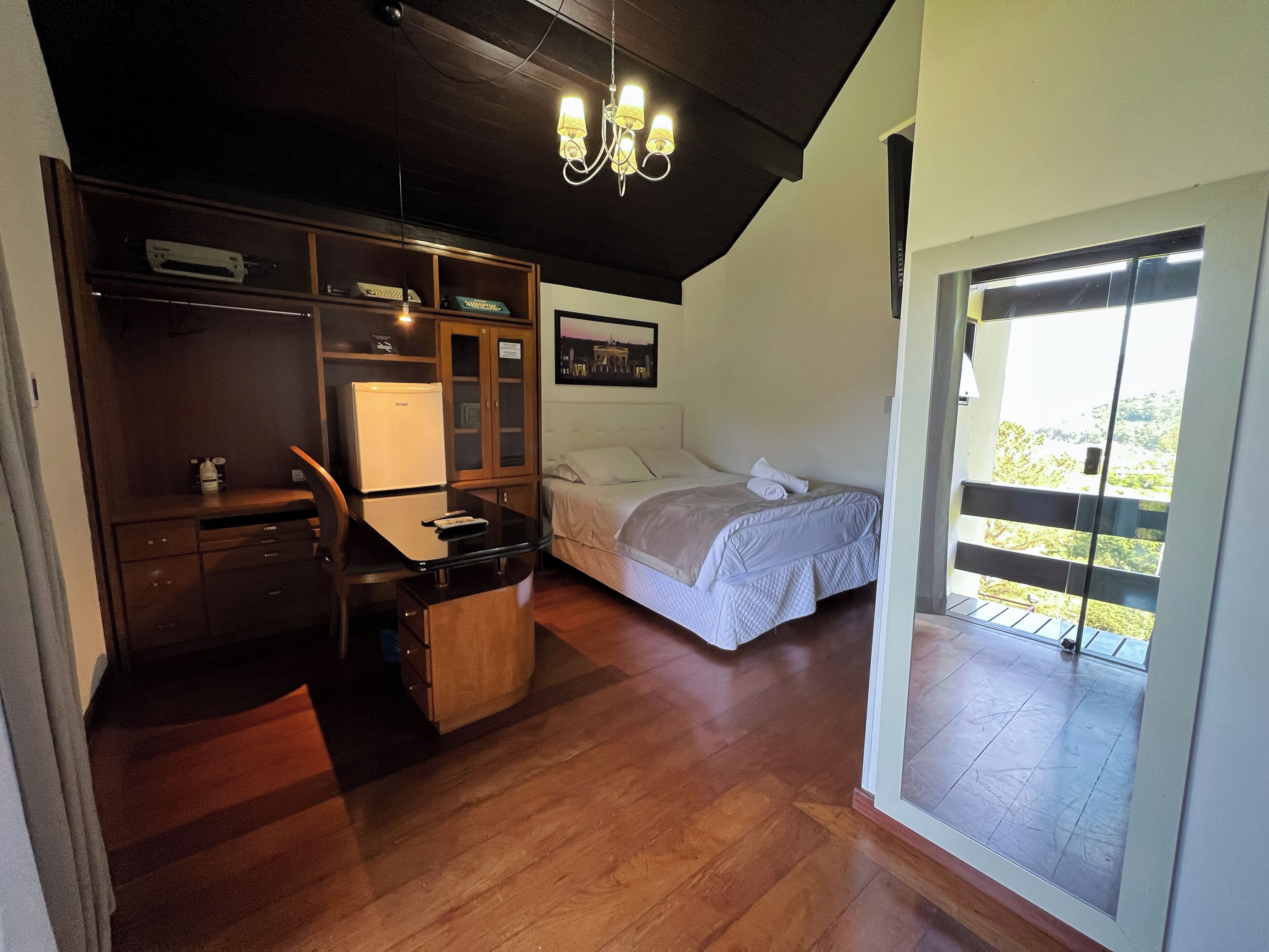 A bedroom with a bed, a desk, a refrigerator, and a view of the outdoors.
