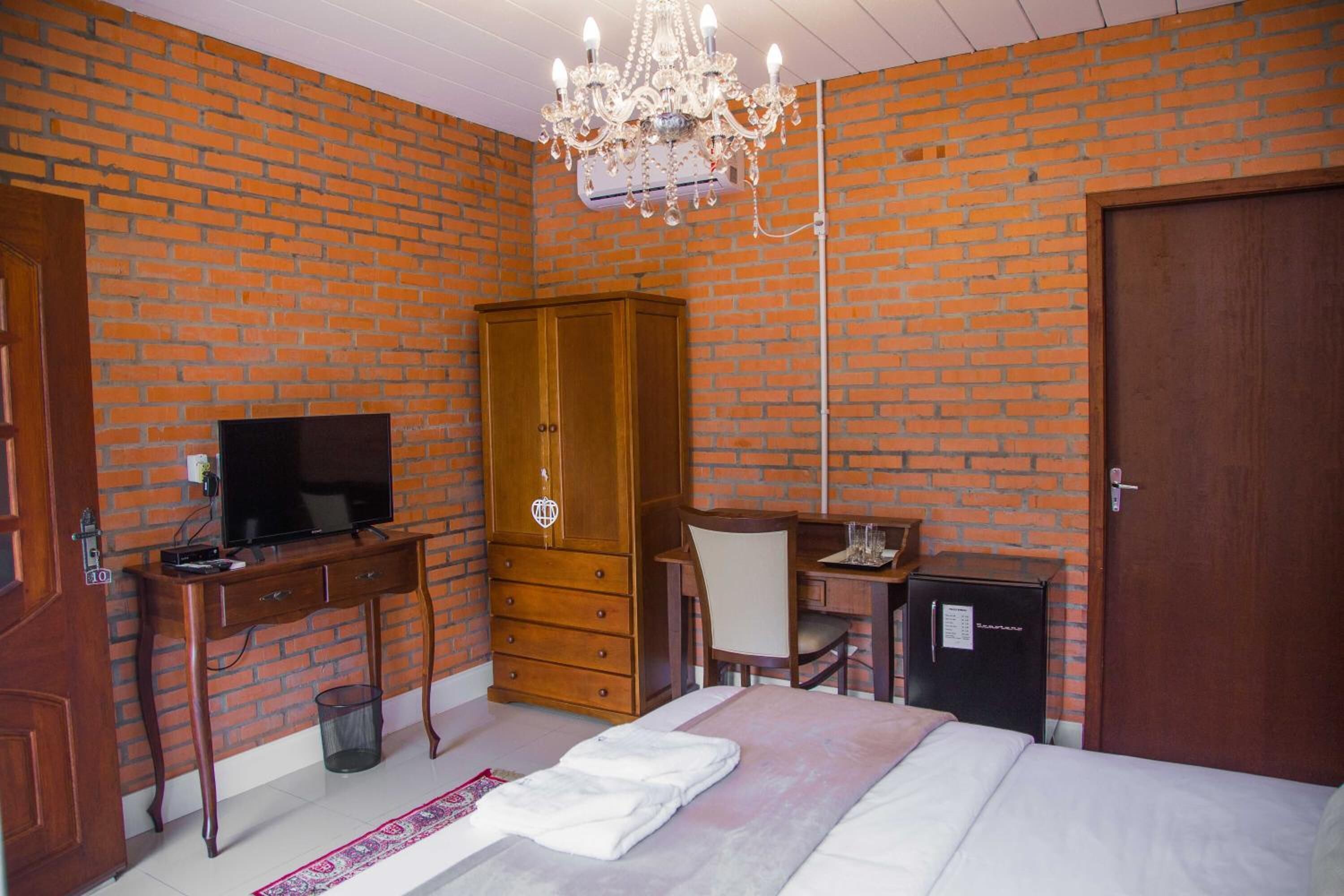 Ein Hotelzimmer mit Backsteinwand, Holzschrank, Schreibtisch mit Stuhl, Fernseher, Bett und kleinem Kühlschrank.