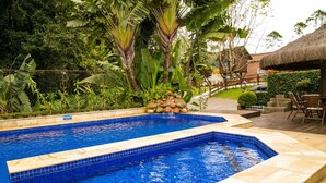 Una piscina con borde de azulejos, rodeada de exuberante vegetación y una estructura de techo de paja.
