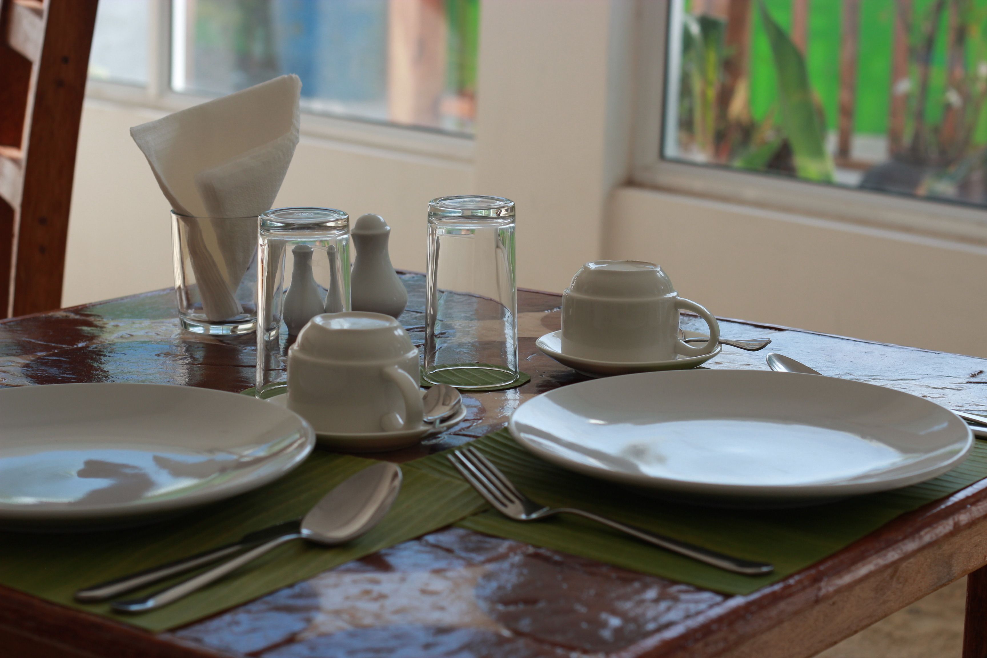 Une table à manger dressée avec des assiettes, des couverts, des verres et une serviette, avec une fenêtre en arrière-plan.