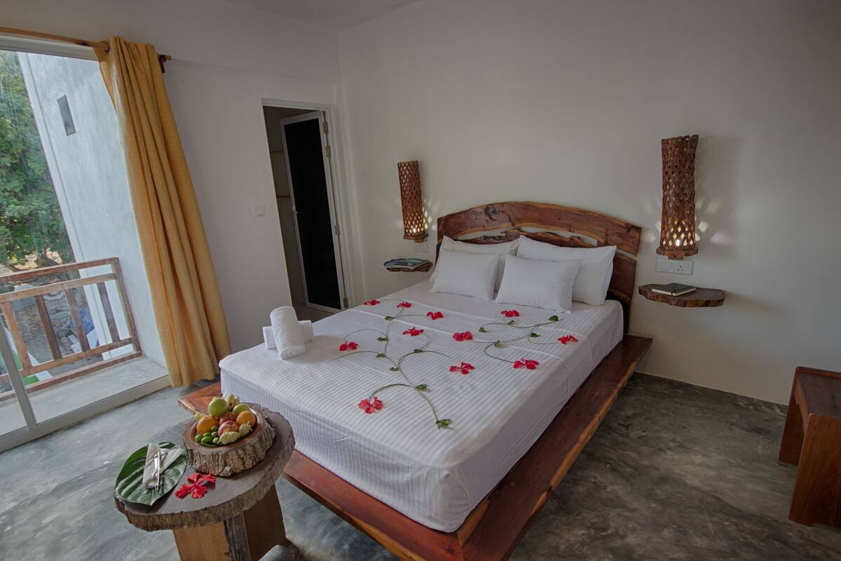 A neatly made bed with a floral bedspread, a bedside table with a fruit bowl, and a view of the outdoors through a balcony door.