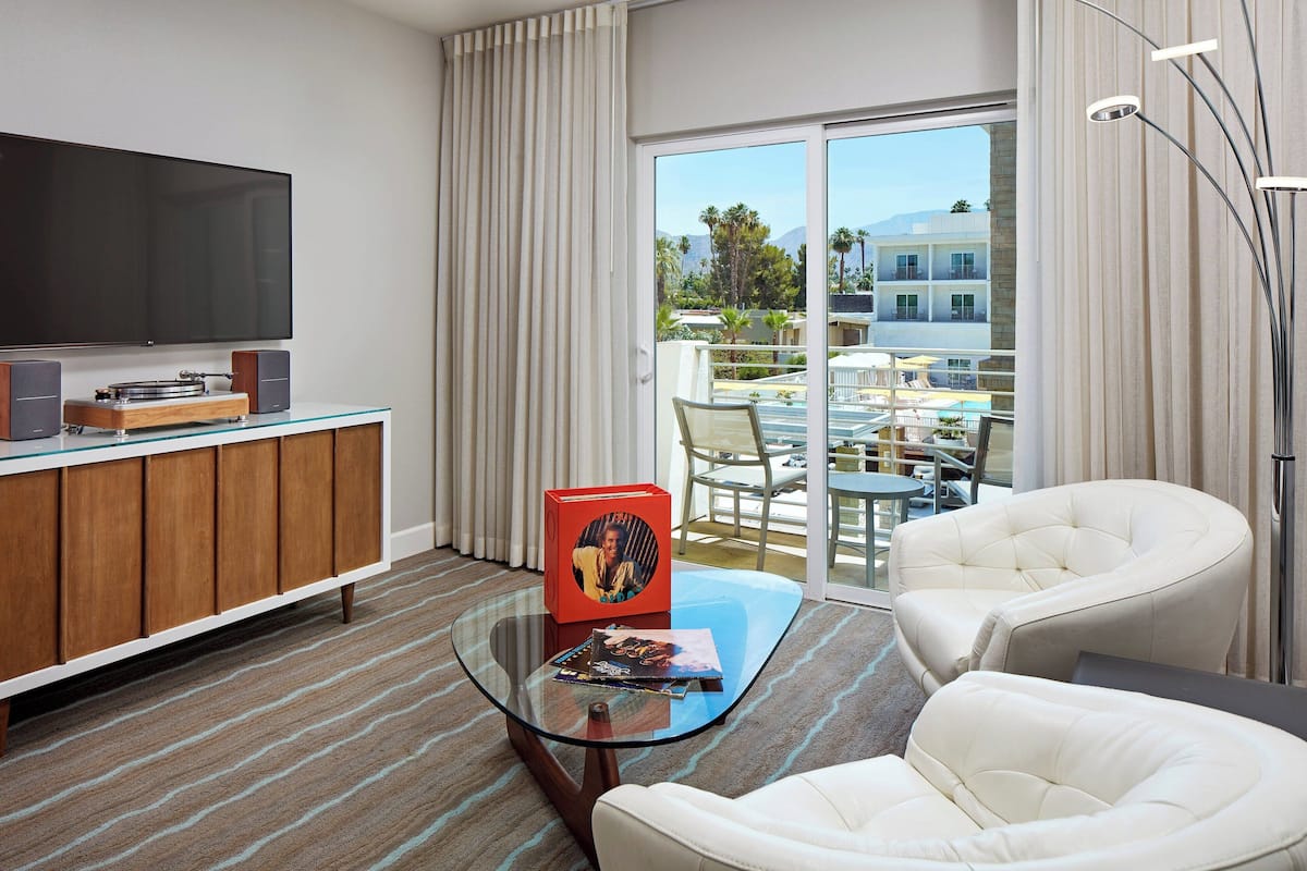 A modern hotel room with a glass coffee table, a white armchair, a flat-screen TV, and a view of the outdoor pool area.