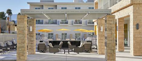 Un espace extérieur aménagé, abrité et doté de mobilier en osier et de parasols jaunes, situé devant un bâtiment moderne avec des balcons.