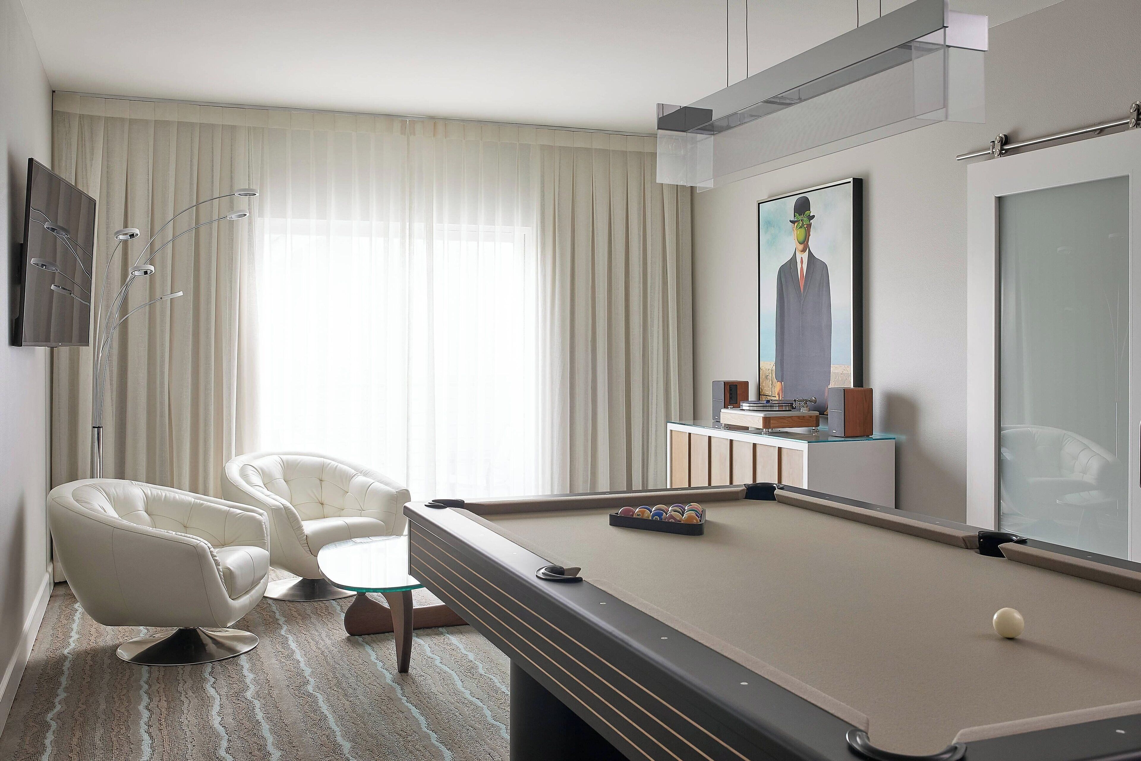 A modern hotel room with a pool table, two white armchairs, a glass-top coffee table, and a large painting on the wall.