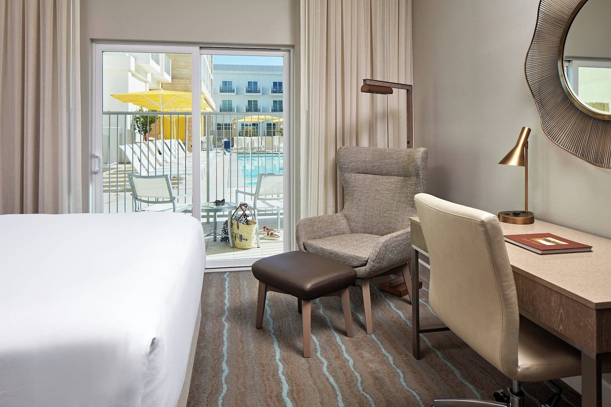 A hotel room with a bed, a desk, a chair, a small stool, a lamp, and a view of a pool and building through a sliding glass door.
