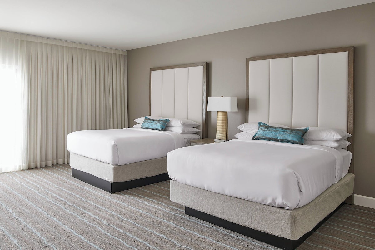 A hotel room with two beds, a large window with curtains, a lamp, and a striped carpet.