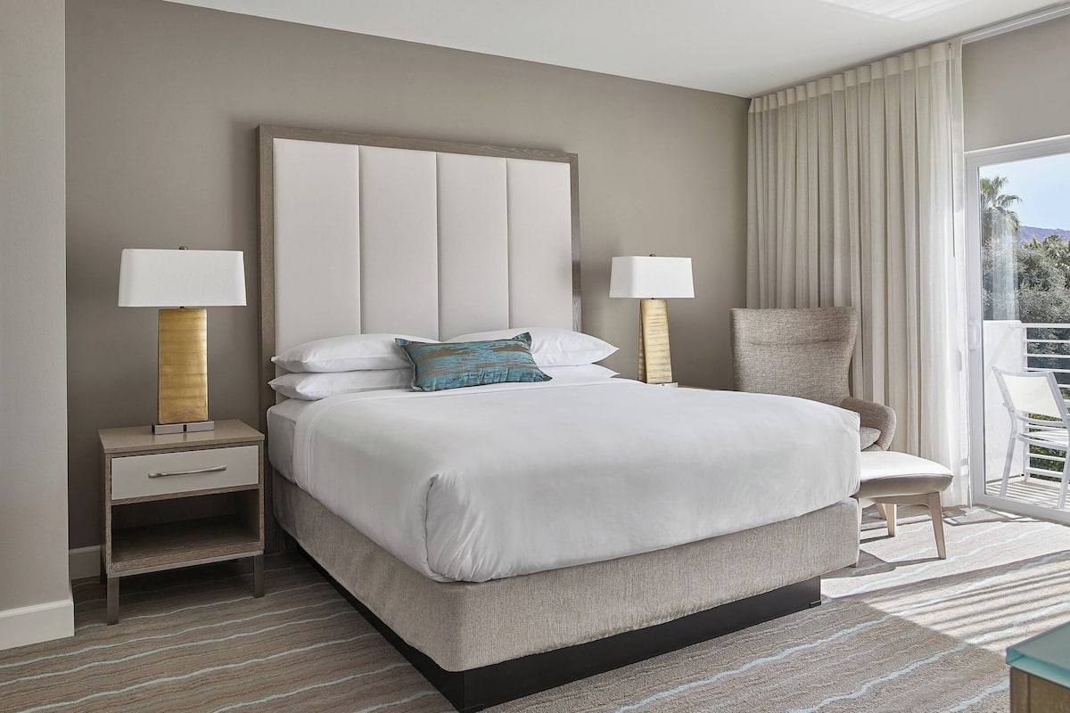 A modern hotel room with a large bed, two bedside tables with lamps, a chair, and a balcony with curtains.