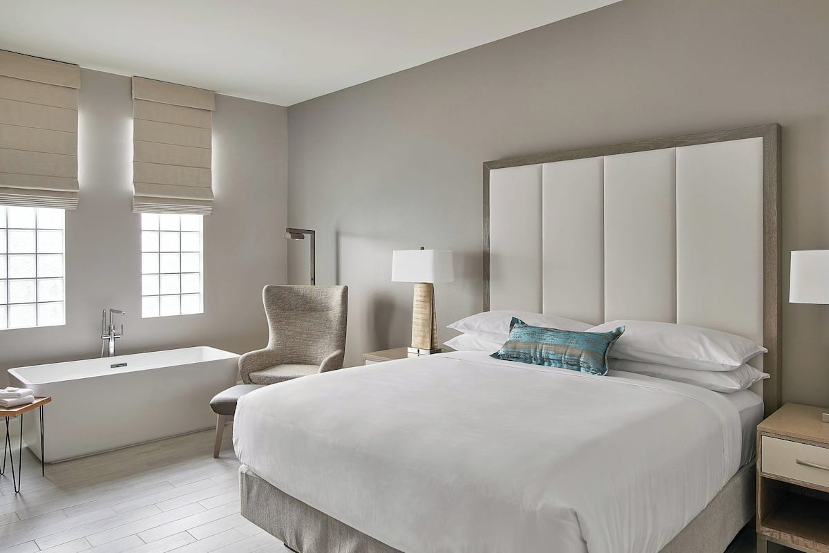 A modern hotel room with a large bed, a sitting area with a chair, a bathtub, and a bedside table with a lamp.