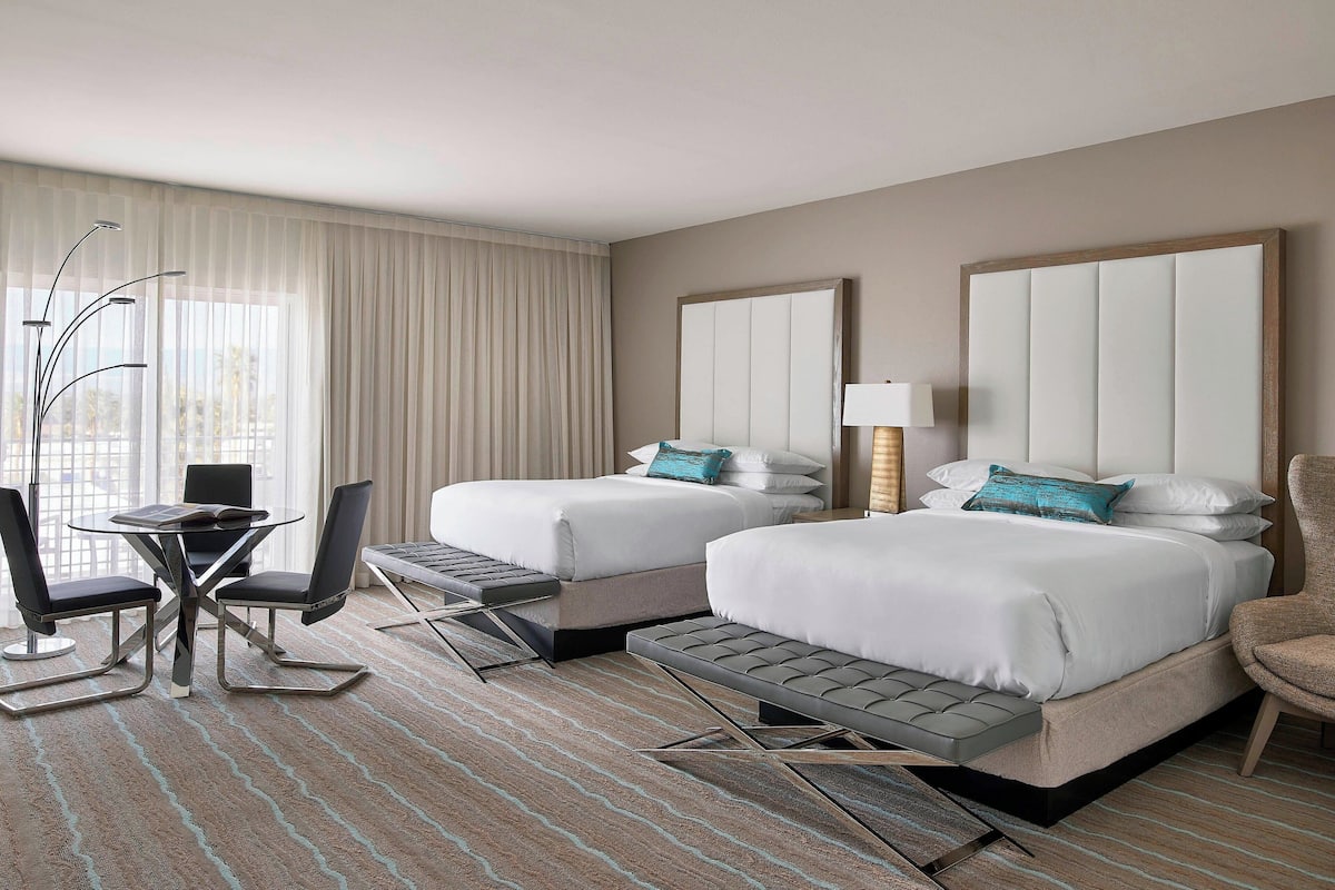 A hotel room with two beds, a desk with chairs, a lamp, and a view of the outdoors.
