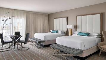 A hotel room with two beds, a desk with chairs, a lamp, and a view of the outdoors.