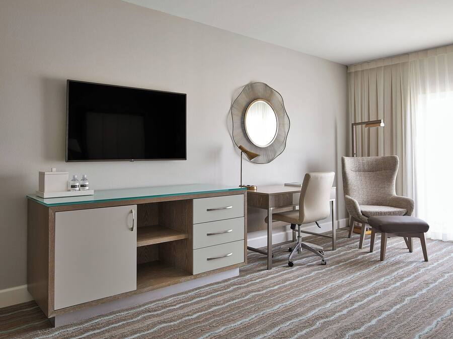 Habitación de hotel con televisor de pantalla plana, espejo redondo, escritorio con silla y ventana grande con cortinas.