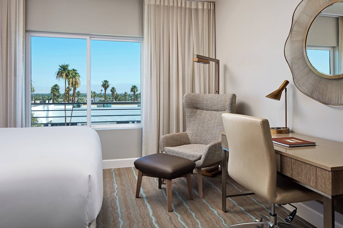 A hotel room with a bed, a desk, a chair, a small stool, a lamp, and a large window offering a view of palm trees and a distant mountain.
