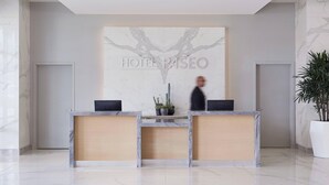 A hotel reception desk with a marble design, a large plant, and a guest walking by. - Hotel Paseo, Autograph Collection by Marriott (Palm Desert)
