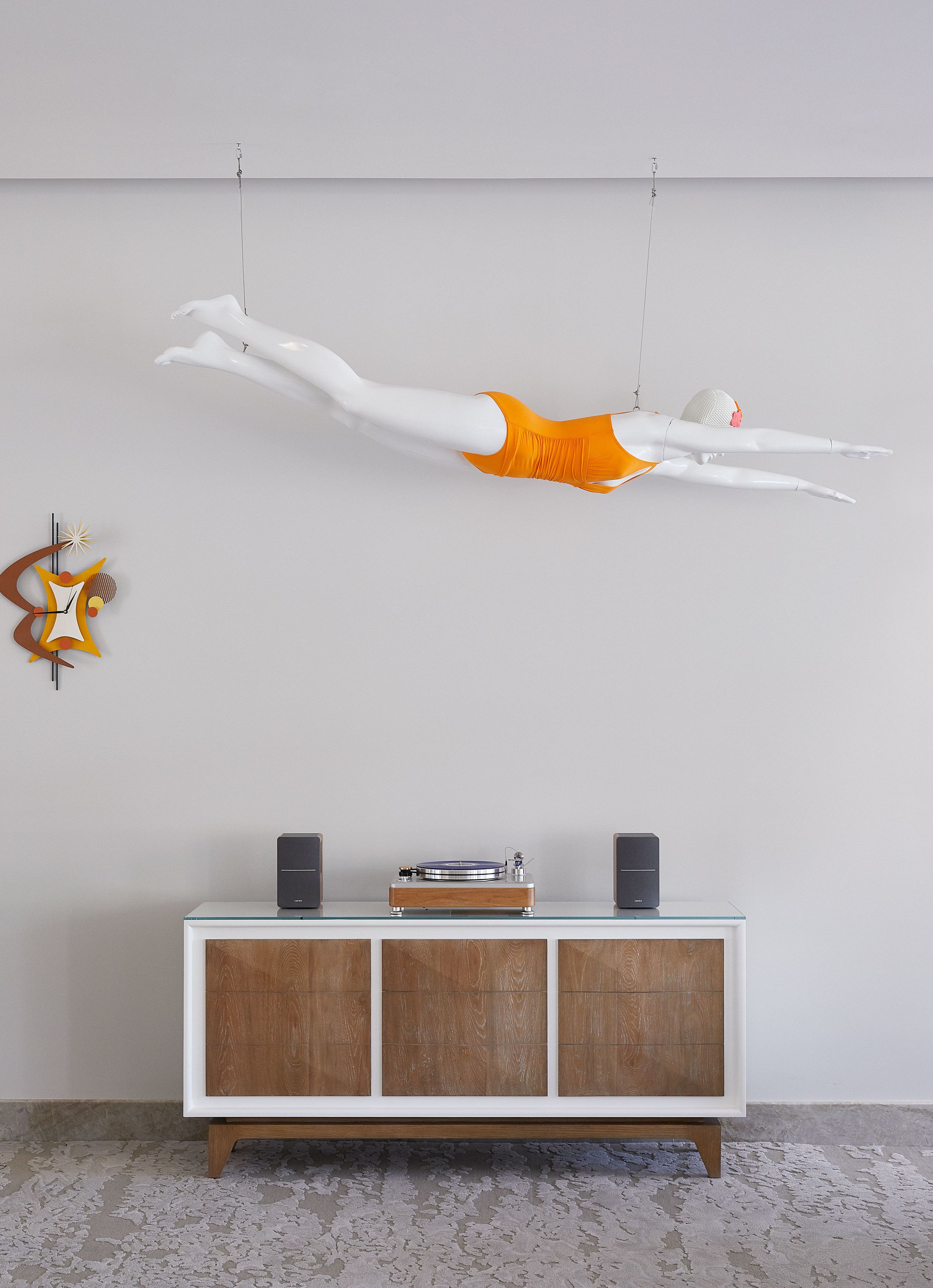 A mannequin in a white and orange outfit suspended from the ceiling, a wooden cabinet with speakers, and a textured floor.