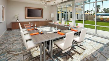 A conference room with a long table, white chairs, and a large window with a view of palm trees and a bus.