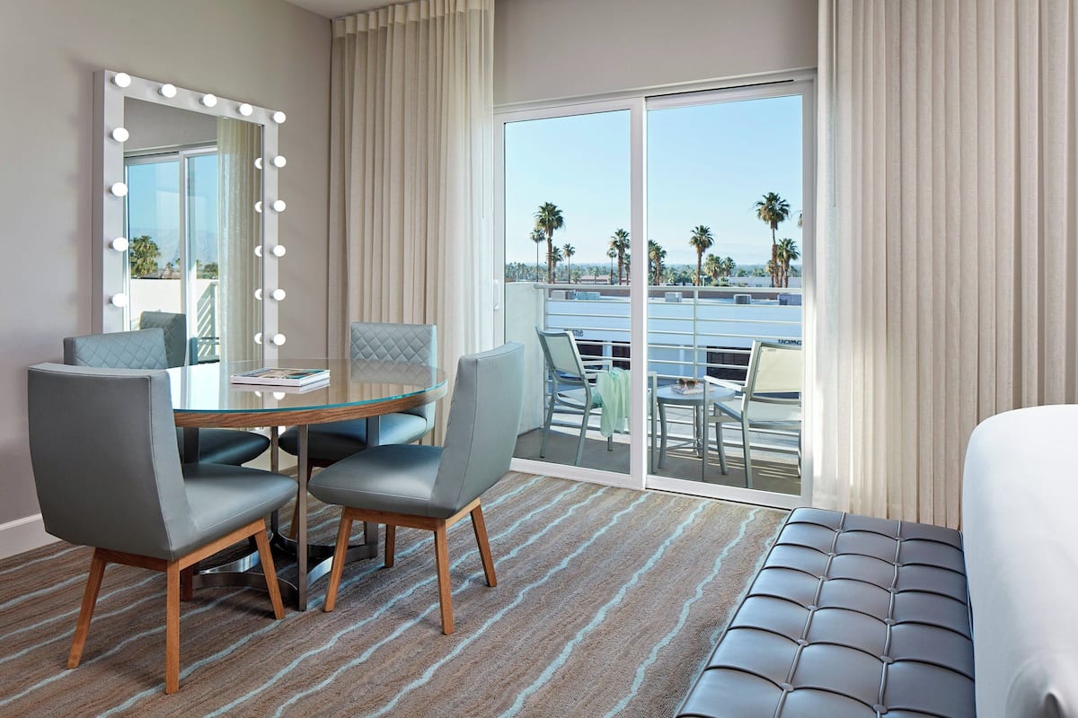 A hotel room with a large mirror with lights, a round glass table, two chairs, a bed, and a view of palm trees and a pool.
